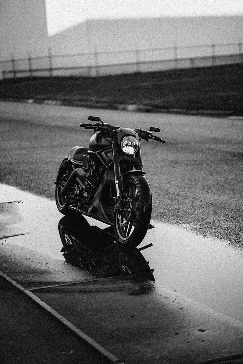 Dark motorcycle reflected in a rain puddle on textured asphalt with a blurred fence and building background.