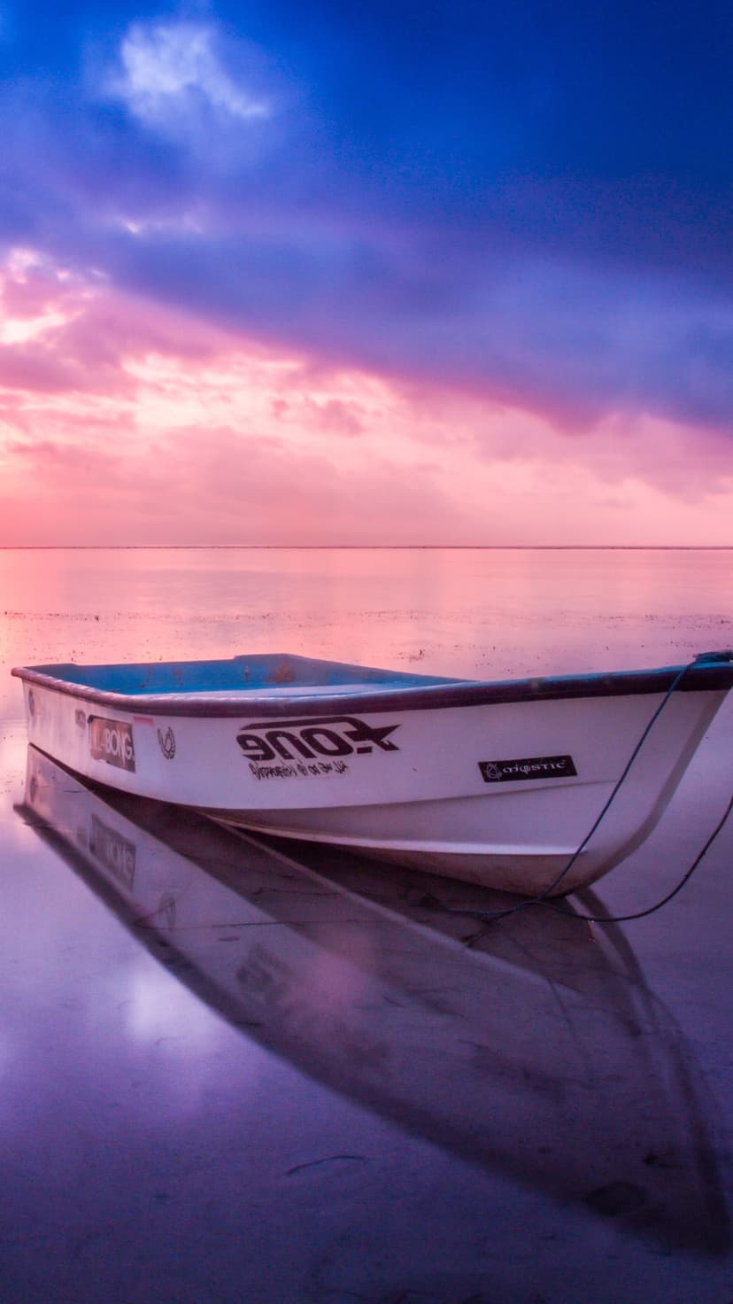 Small white boat floats on calm reflective water under a pink and purple pastel sunrise sky with soft clouds.