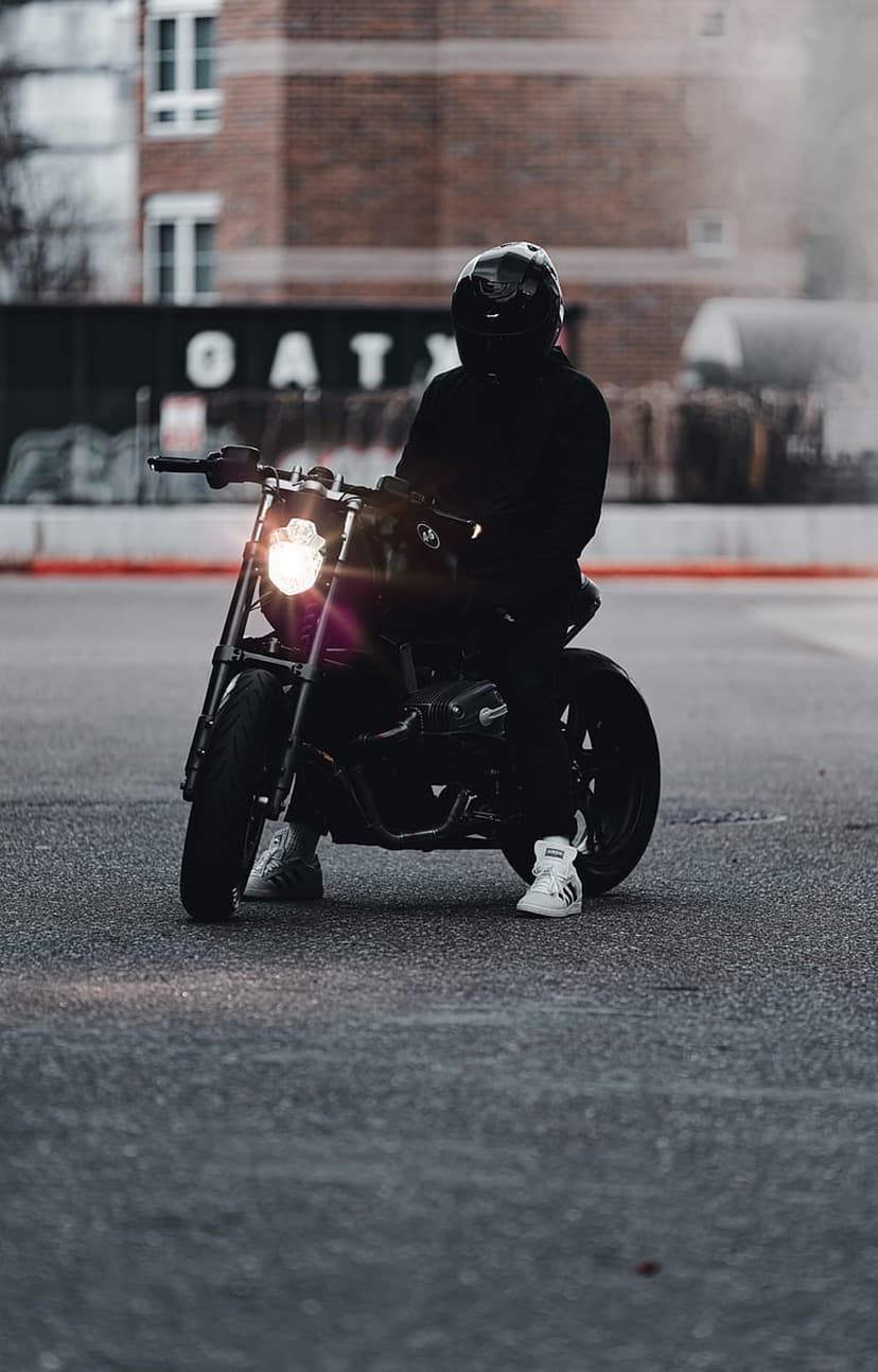 Motorcyclist in a black helmet sits on a dark bike with an illuminated headlight against a blurred brick wall.