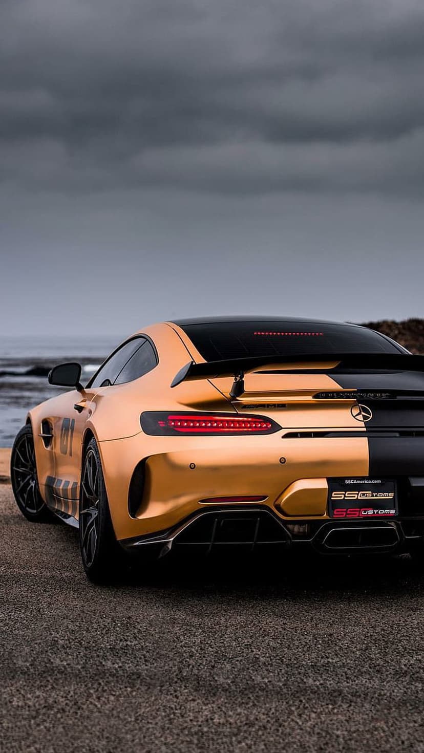Mercedes-AMG GT S with gold and black paint parked on gravel by the ocean under a cloudy sky in a rear view shot.