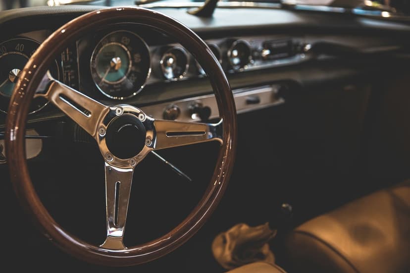 Polished wood steering wheel with chrome spokes and vintage analog gauges inside a classic car's leather interior.