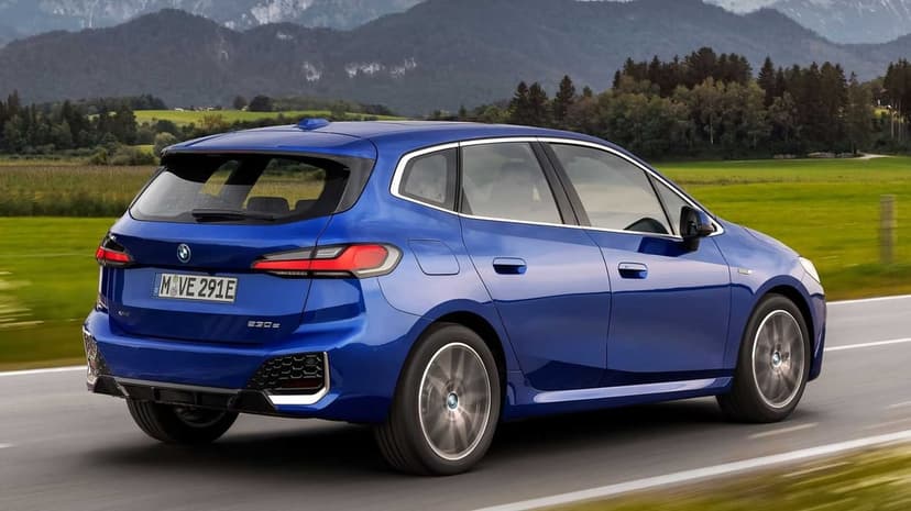Rear view of a blue BMW 230e driving on a road with misty mountains and green fields in the background.