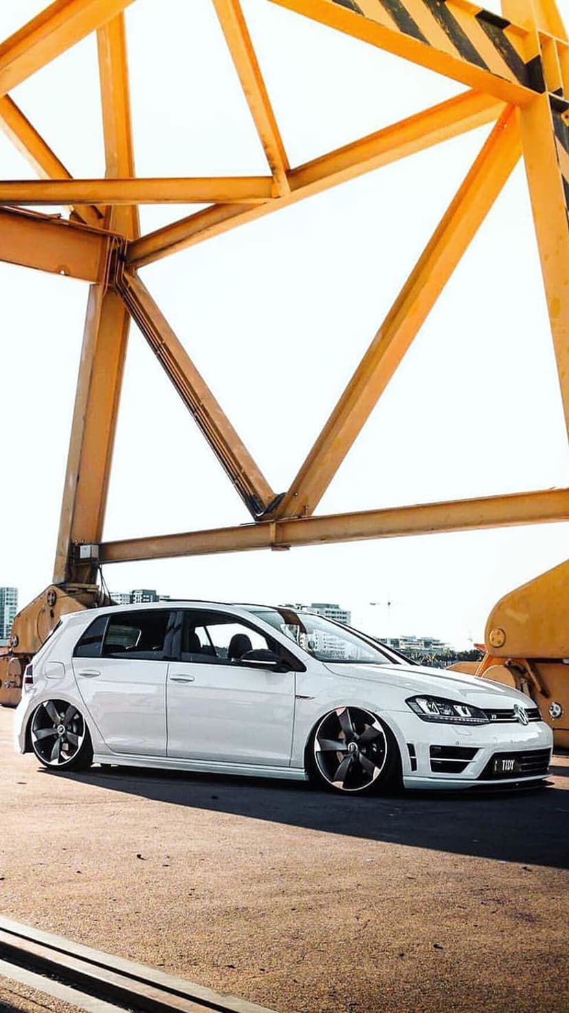 White Volkswagen Golf R parked under a massive yellow crane with a low angle view and bright urban background.