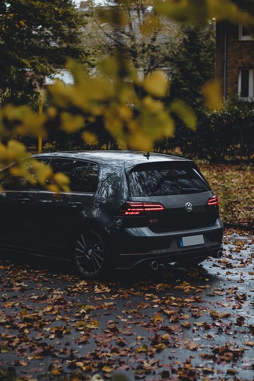 Dark Volkswagen Golf GTI parked on autumn leaves with glowing red taillights on a damp street at twilight.