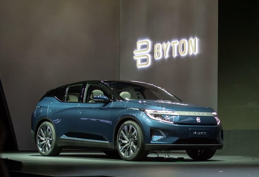 Metallic blue BYTON M-Byte electric SUV on an exhibition stage with a glowing brand logo in the background.