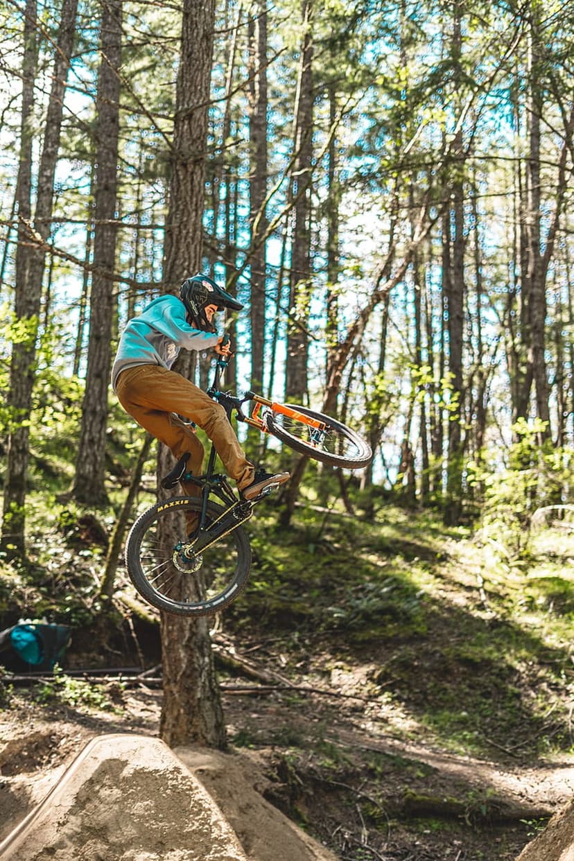 Mountain biker suspended mid-air during a dirt jump on a sun-drenched forest trail with tall evergreen trees.