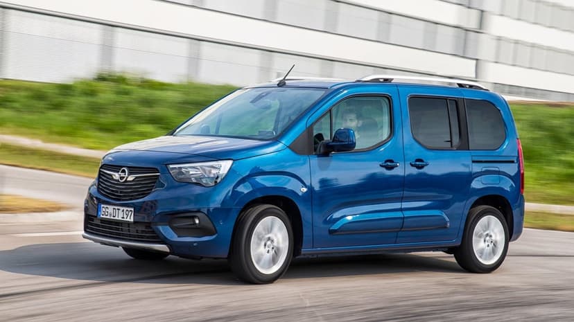 Blue Opel Combo Life driving on a paved road with motion blur on wheels and a modern building background.