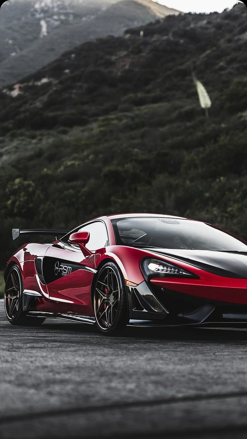 Red McLaren supercar with black accents parked on a winding asphalt road against dark green mountain hills.