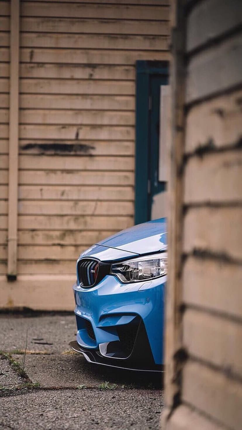Blue BMW M4 front fascia with sharp headlights parked against a weathered vertical wooden plank wall.