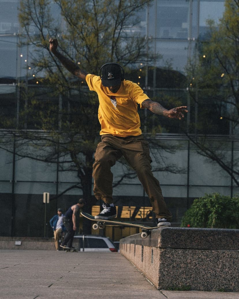 Skateboarder in a yellow shirt grinds a concrete ledge in front of a glass building and green trees in daylight.
