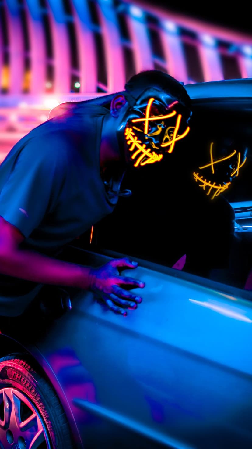 Masked person with glowing blue eyes leans against a dark car amidst vibrant neon purple and orange city lights.