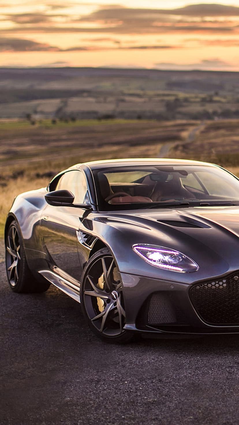 Aston Martin DBS Superleggera with glowing headlights on a rural road during a golden sunset vista.