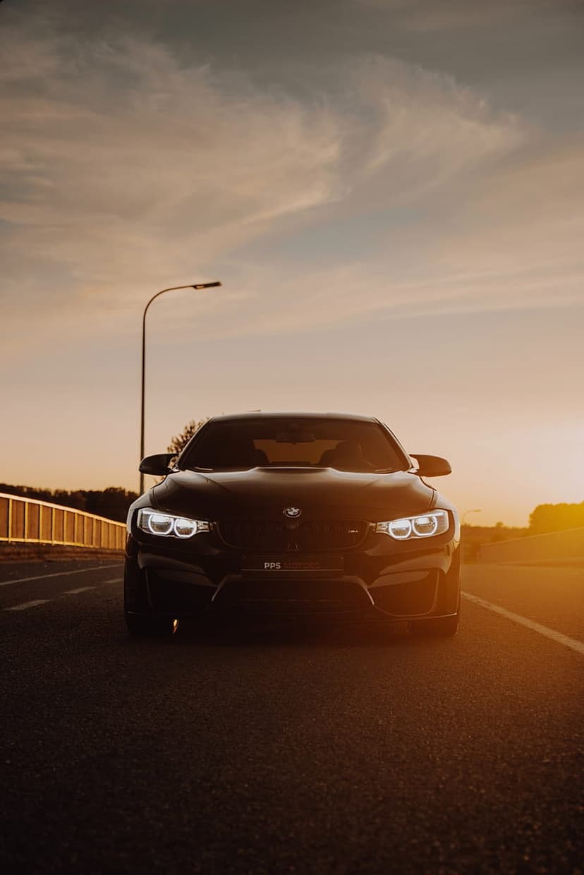 Black BMW M4 sports car parked on an asphalt road at sunset with glowing headlights and golden sky reflections.