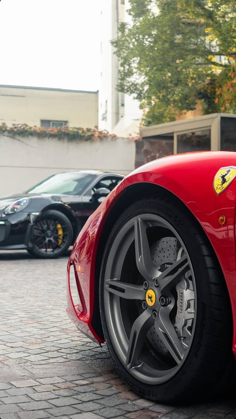 Red Ferrari wheel and fender in focus with a black Porsche parked on cobblestones under bright sunlight.