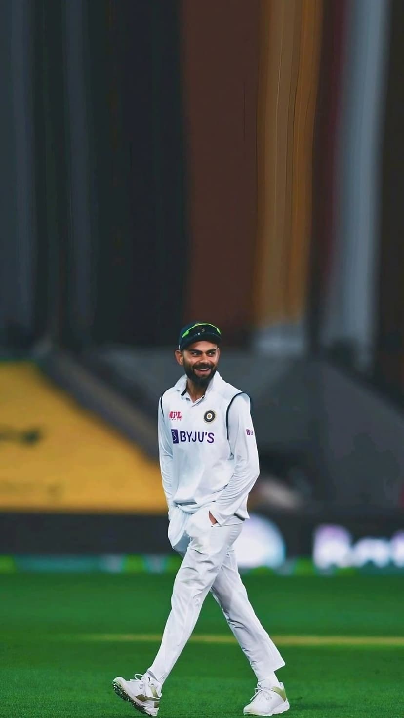 Virat Kohli smiles while wearing a white Indian Test jersey and green cap on a cricket field with stadium seating.