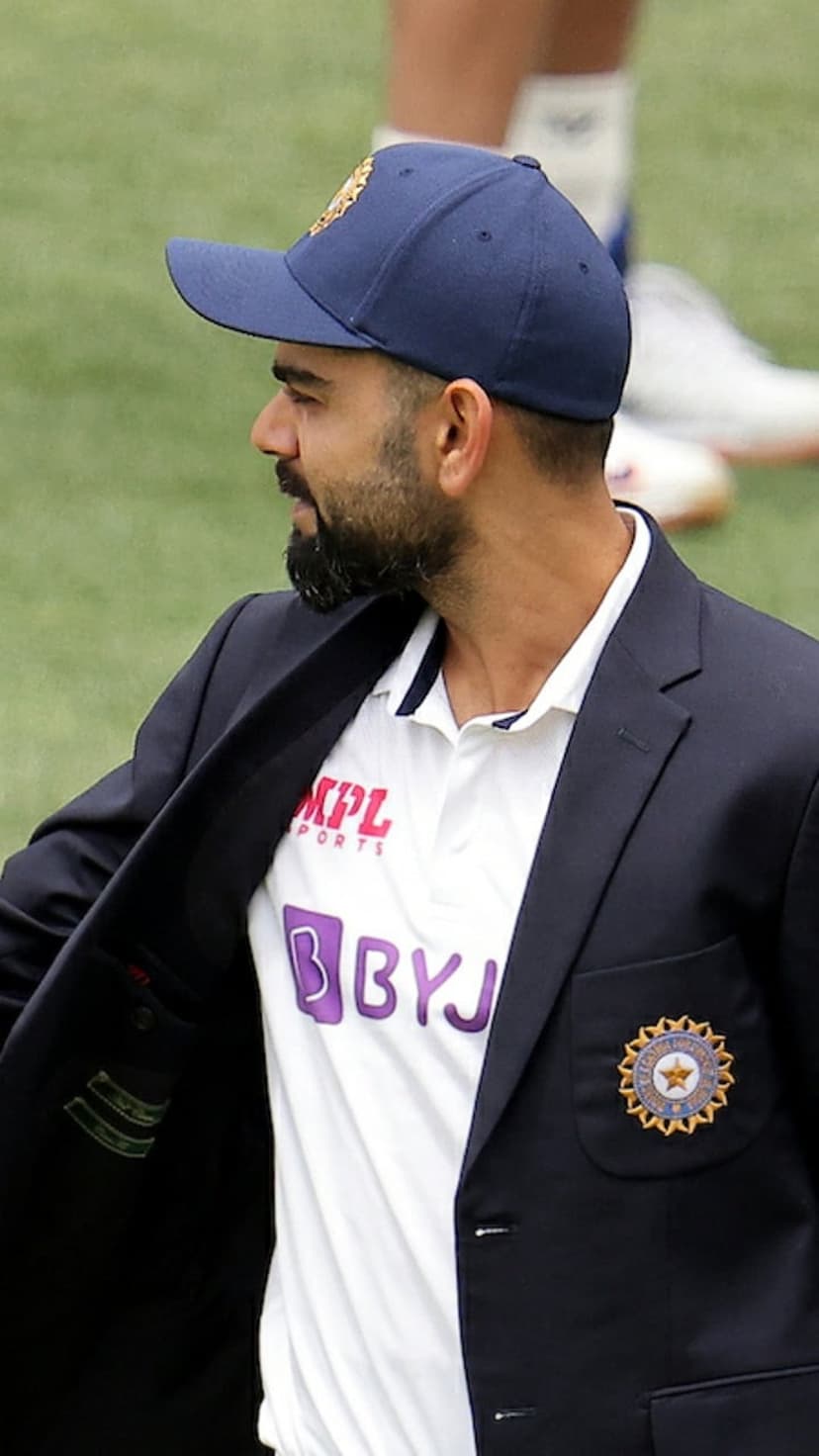 Virat Kohli in a navy blue Indian cricket team blazer and cap, shown in a close-up profile view with BCCI insignia.