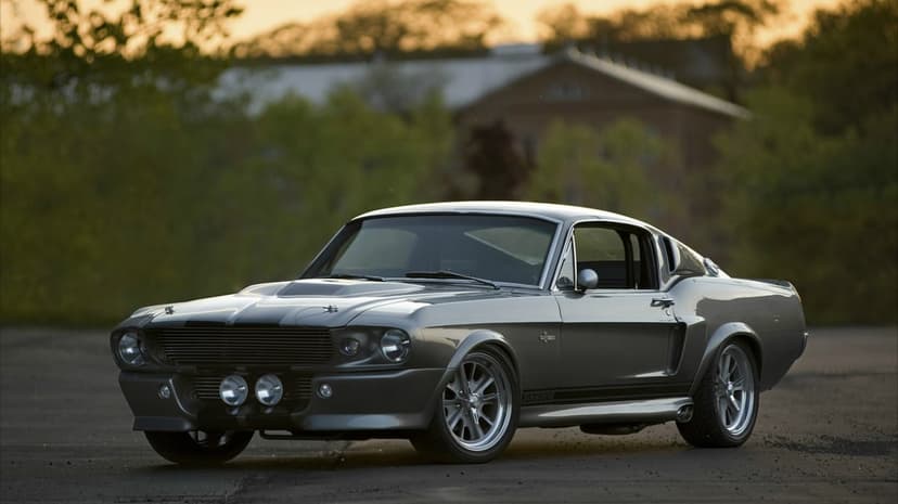 Silver Shelby GT500 Mustang with black stripes parked on asphalt during a warm sunset with blurred trees.