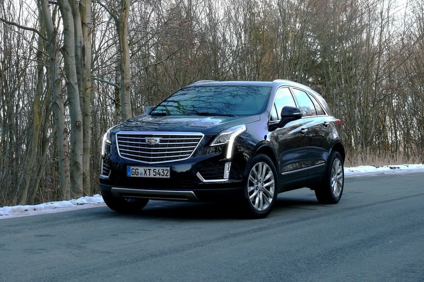 Black Cadillac XT5 SUV parked on a paved road through a forest with bare trees and patches of roadside snow.