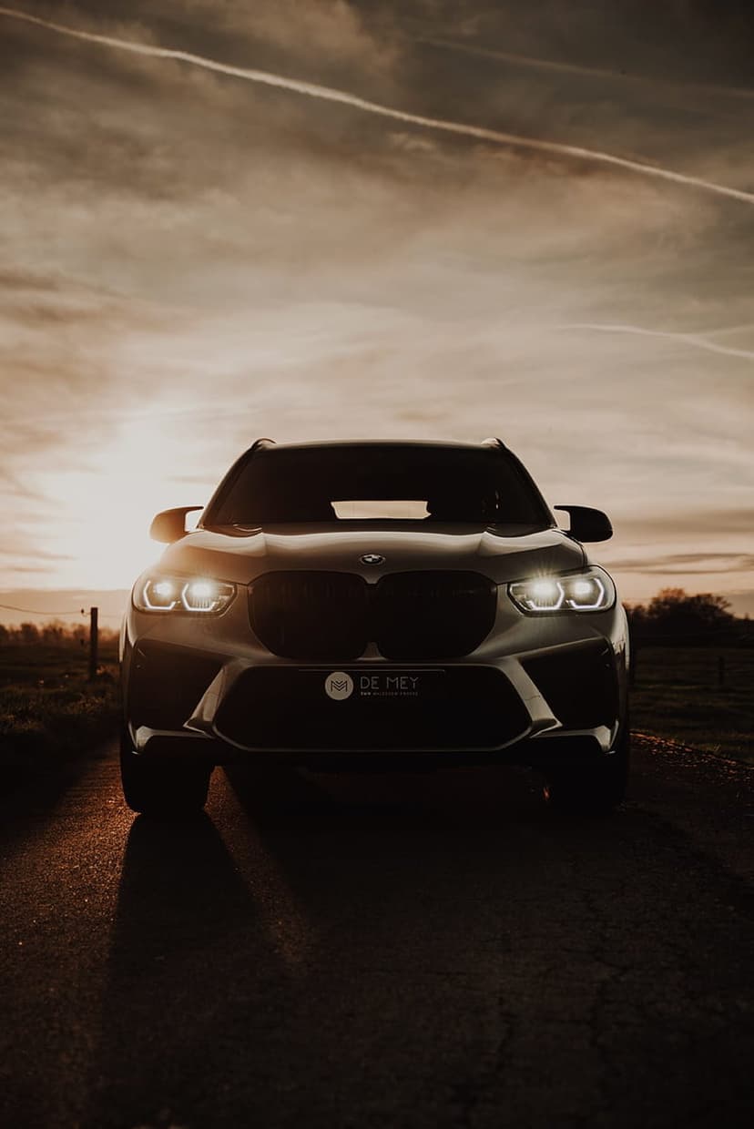BMW X4 M Competition SUV with glowing headlights parked on a rural road under a vibrant golden sunset sky.