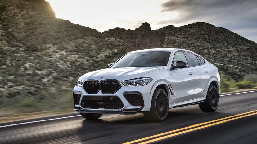 White BMW X6 M SUV driving on a mountain road with sunset light hitting rugged peaks in the background.