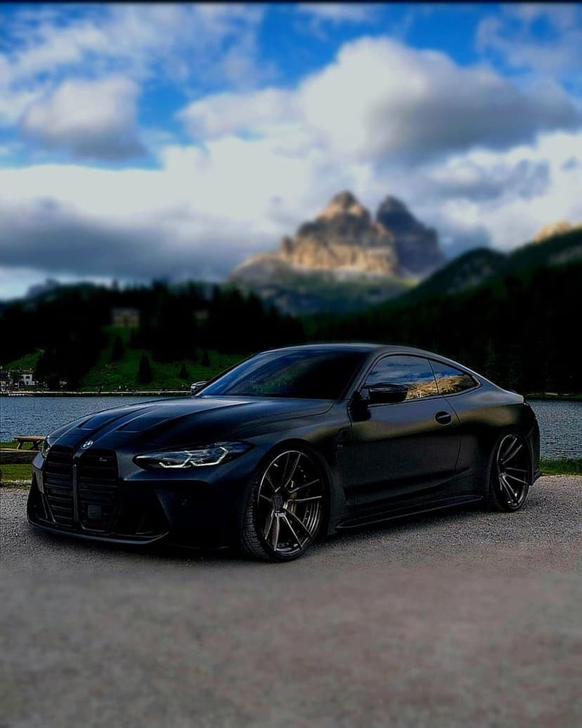 Black BMW M4 coupe parked on a grey gravel road near a calm mountain lake under a moody, overcast sky.