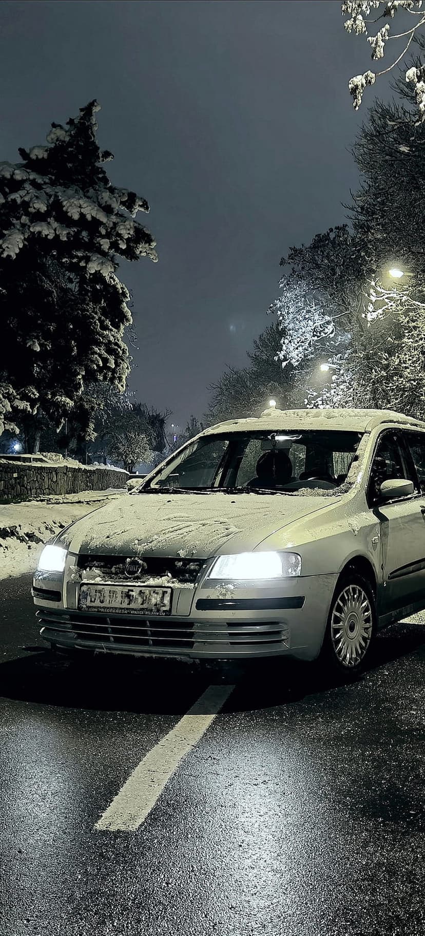 Silver car with glowing headlights parked on a wet asphalt road lined with snow-covered trees and streetlights.