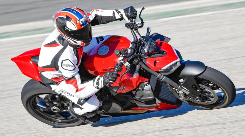 Red Ducati motorcycle and rider in a white racing suit leaning deeply into a curve on a sunny asphalt racetrack.