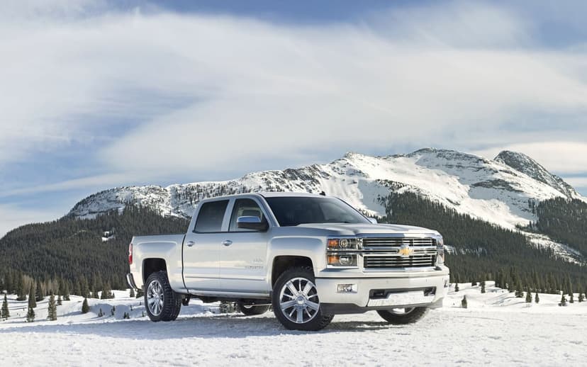 Silver pickup truck on snow with towering white mountains and a blue sky in the background under bright sun.