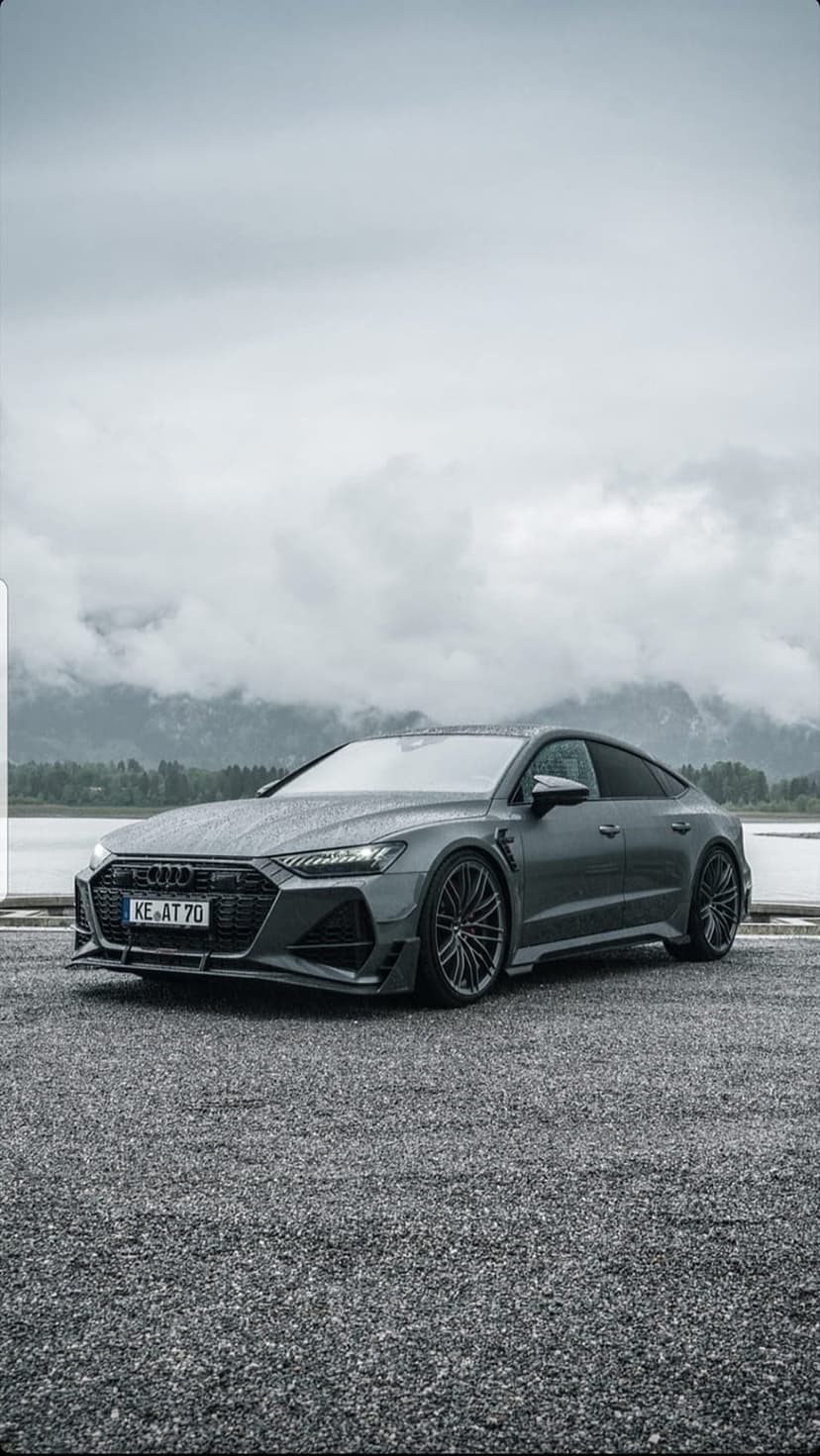 Modified Audi RS7 parked on gravel by a mountain lake under cloudy skies, shown in a vertical mobile orientation.