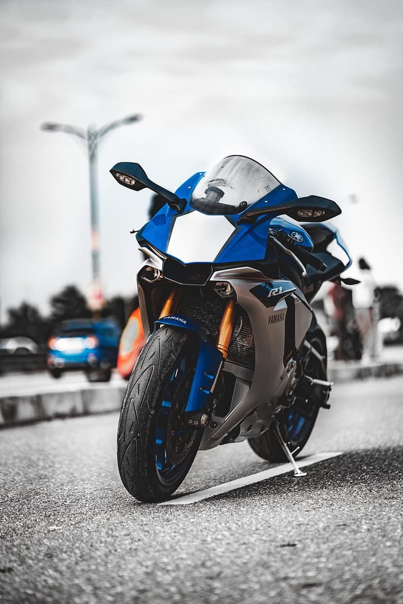 Blue Yamaha R1 sportbike with gold front forks parked on a textured asphalt road with a blurred urban backdrop.