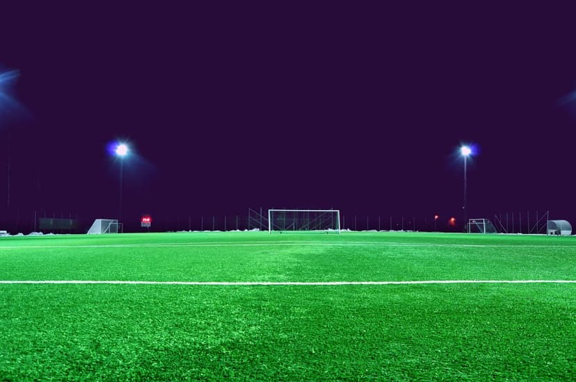 Illuminated soccer pitch with green turf and goalposts under a purple night sky and bright stadium floodlights.