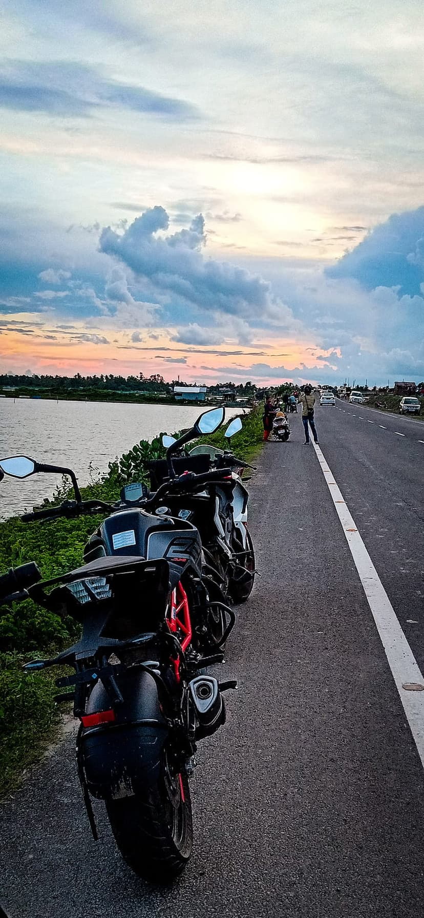 Motorbikes parked beside a winding river under a dramatic sunset sky with orange clouds and calm water.