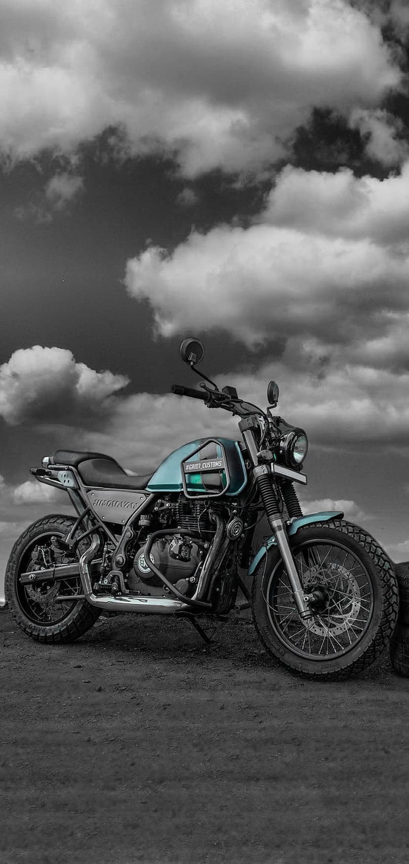 Custom scrambler motorcycle with teal accents parked on jagged rocks beneath a dark and heavy cloud-filled sky.