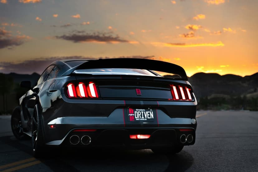 Black Ford Mustang GT with glowing red taillights parked on a highway during a dramatic mountain sunset.