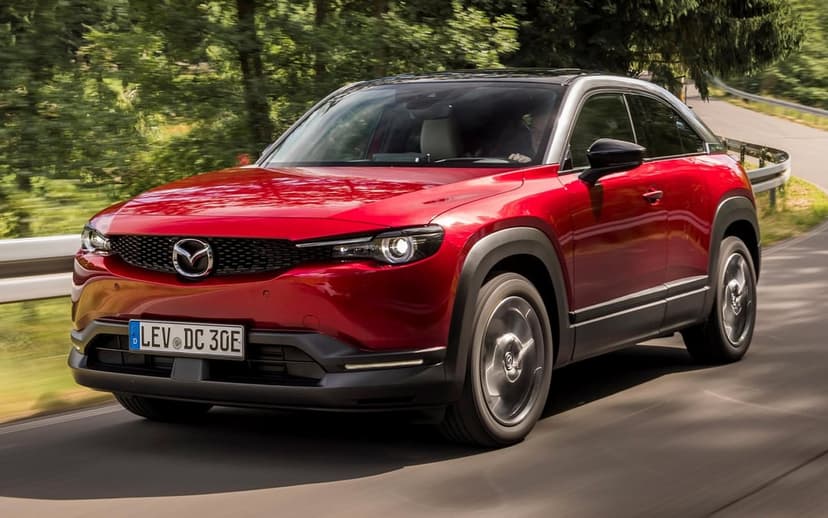Red Mazda MX-30 electric SUV driving along a winding forest road with lush greenery and soft sunlight.
