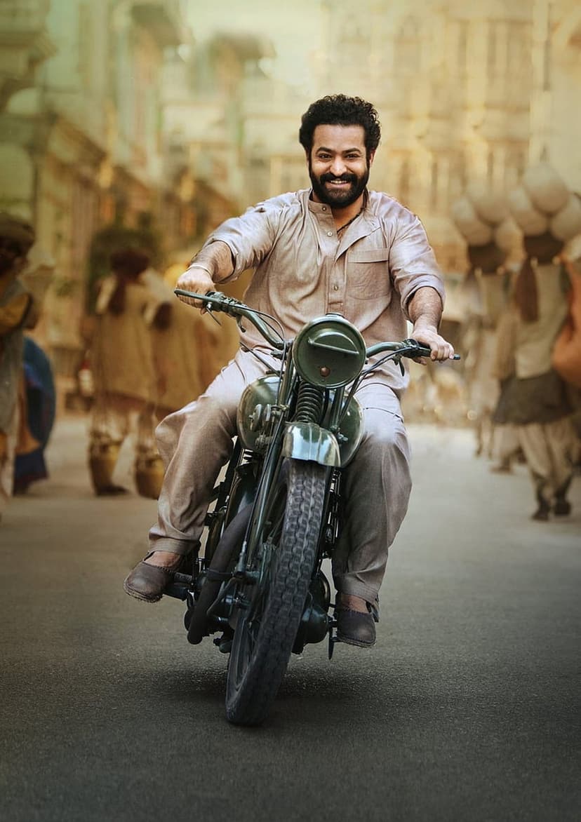 Bearded man in a light kurta smiling while riding a vintage motorcycle on a busy, sunlit Indian village street.
