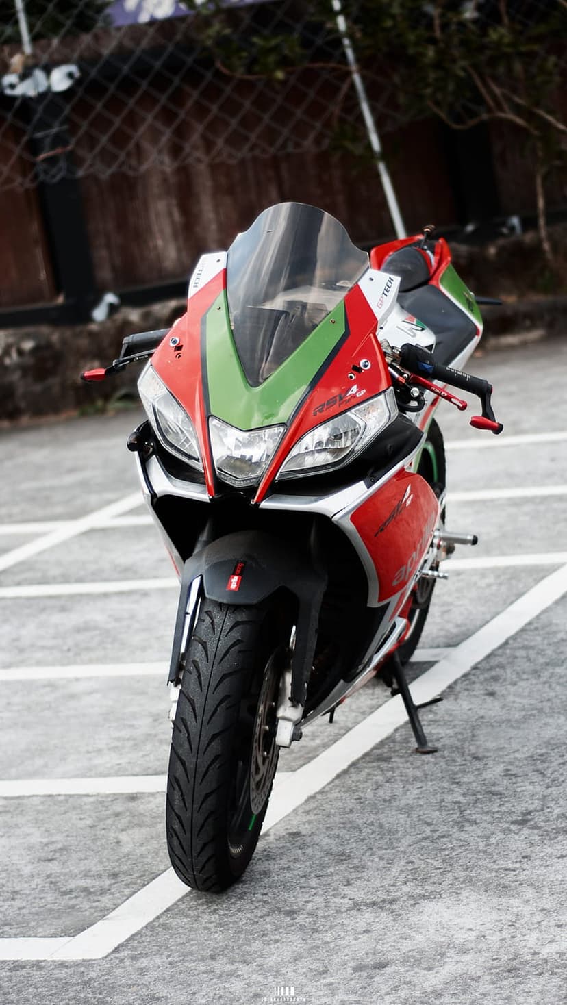 Aprilia RSV4 motorcycle with red and green fairings parked on asphalt facing forward with clear headlights.