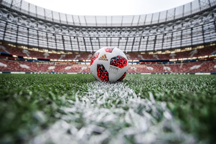 Soccer ball with red patterns on green stadium grass next to a white line with blurred grandstands background.