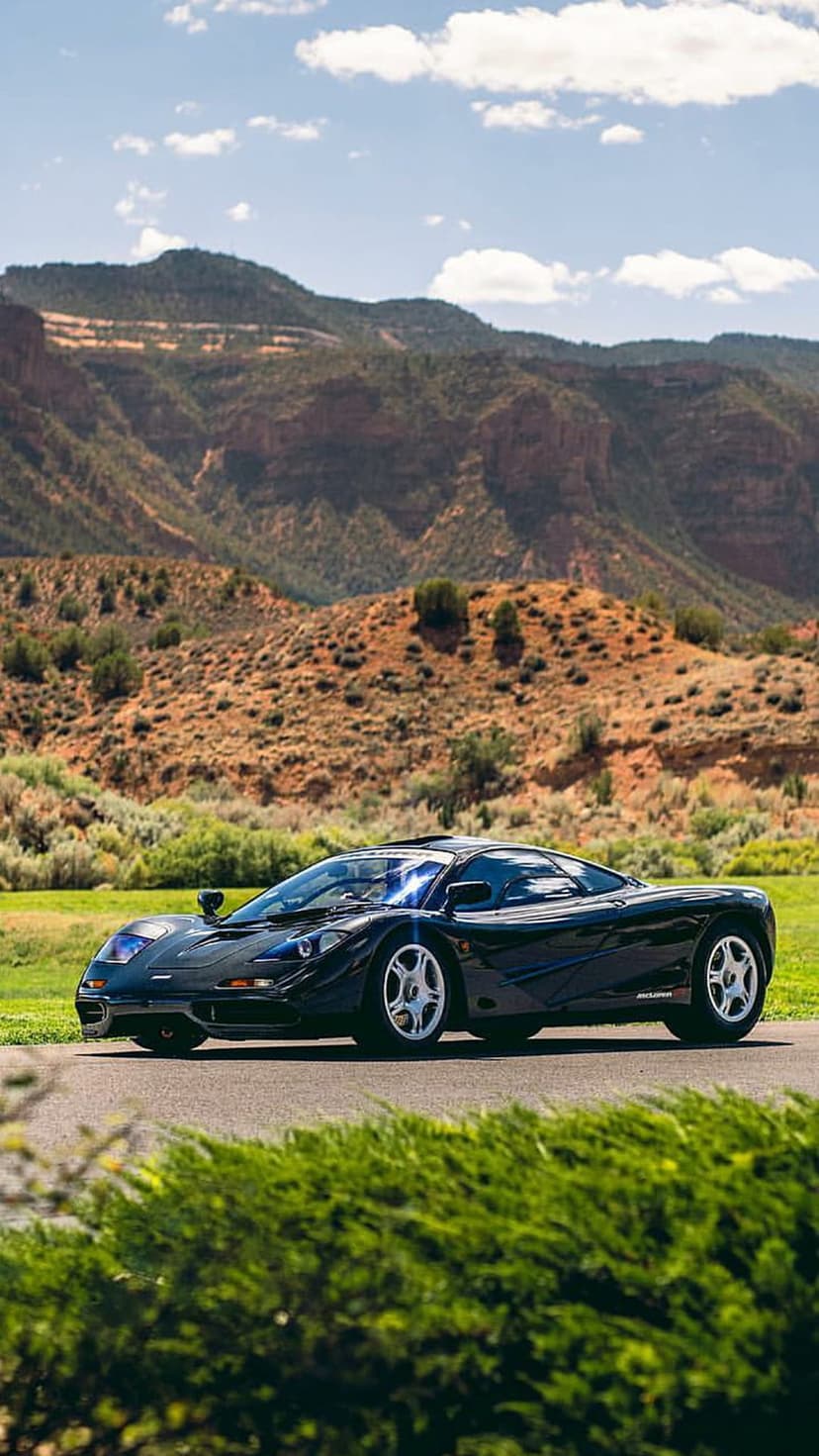 Black McLaren F1 supercar parked on an asphalt road before towering mountains under a bright blue sky.