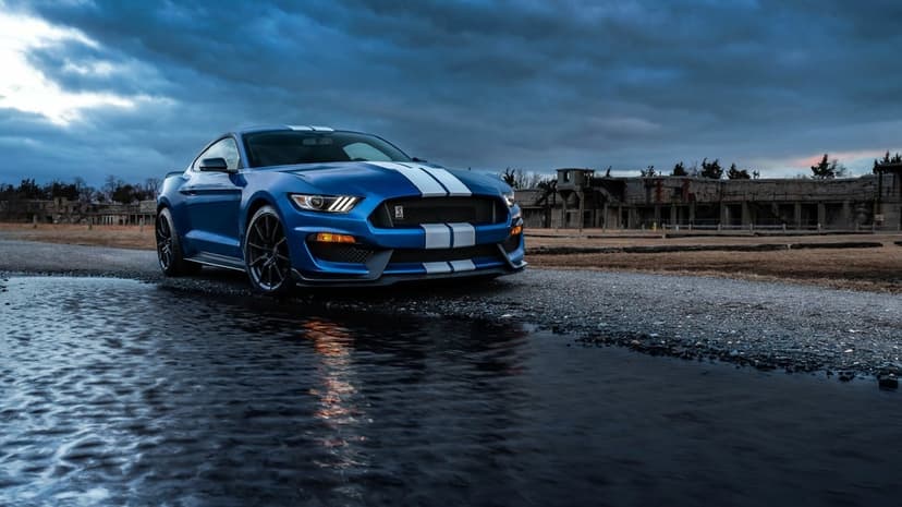 Blue Ford Mustang Shelby GT350R with white stripes on a wet gravel path under a dramatic overcast sky.