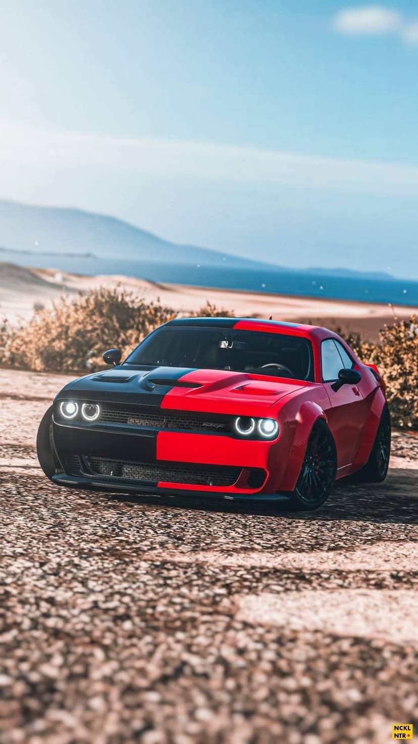 Two-tone red and black Dodge Challenger with glowing ring headlights on a gravel road with a coastal backdrop.