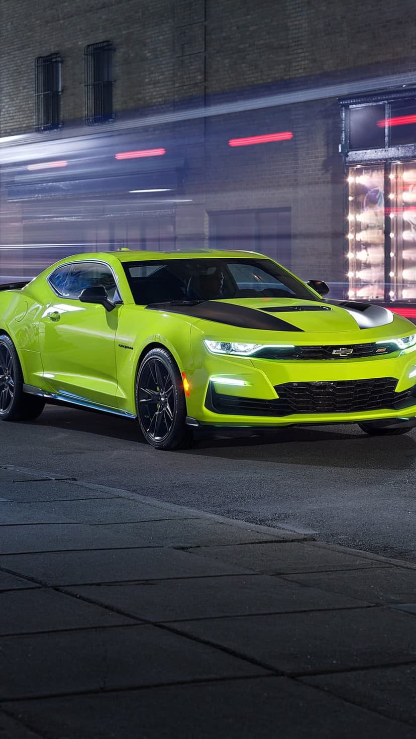 Green Chevrolet Camaro with black hood racing on a night city street featuring glowing LED lights and motion blur.