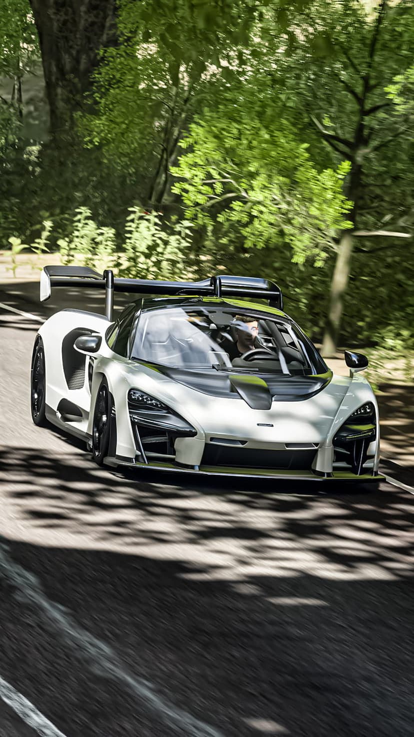 White McLaren Senna supercar accelerating on a forest road with motion blur and sun-dappled light on the asphalt.