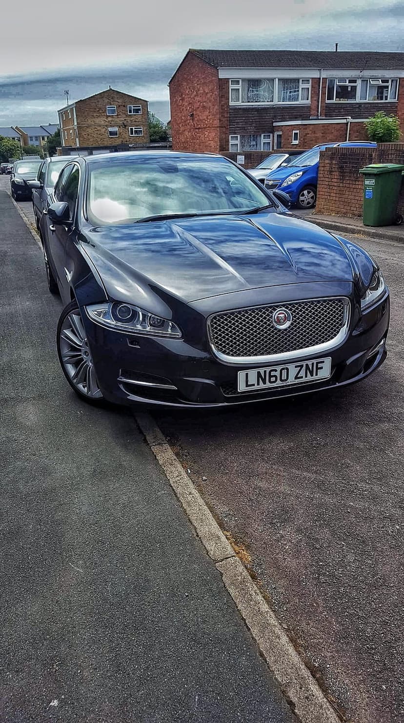 Black Jaguar XJ sedan parked beside a brick house on a cloudy day with a blue car visible in the background.