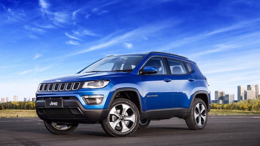 Blue Jeep Compass SUV parked on asphalt under a bright sky with a distant city skyline and trees.