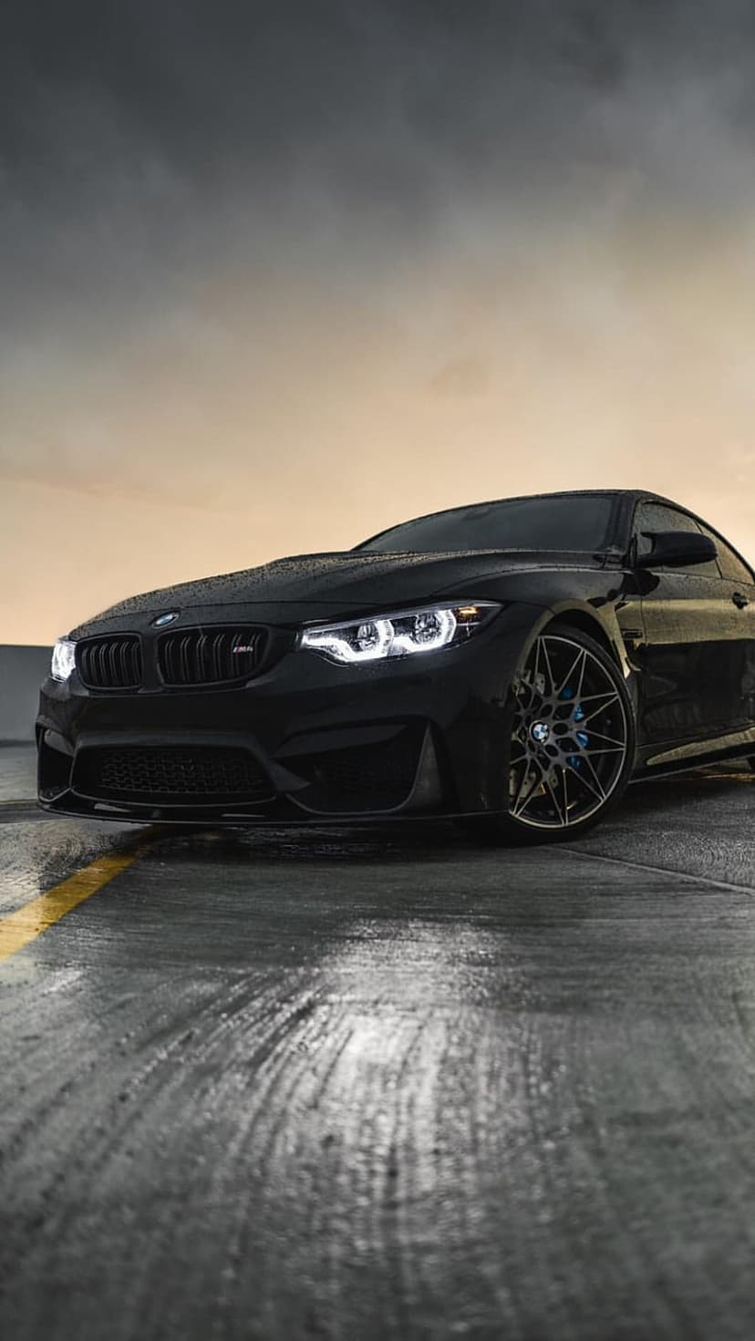 Black BMW M4 coupe with lit headlights on a wet road with yellow lines under a cloudy dusk sky during twilight.