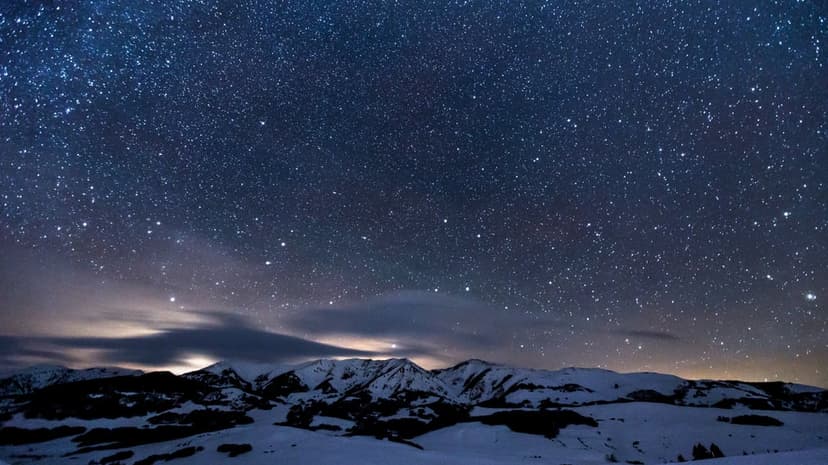 Snow-capped mountain peaks glow under a dense field of stars and drifting clouds during a clear winter night.