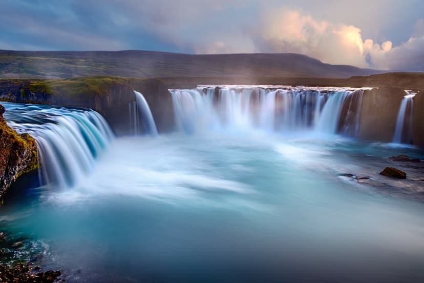 Waterfall cascading into a turquoise pool from a rocky cliff under a cloudy sky in a vertical nature shot.