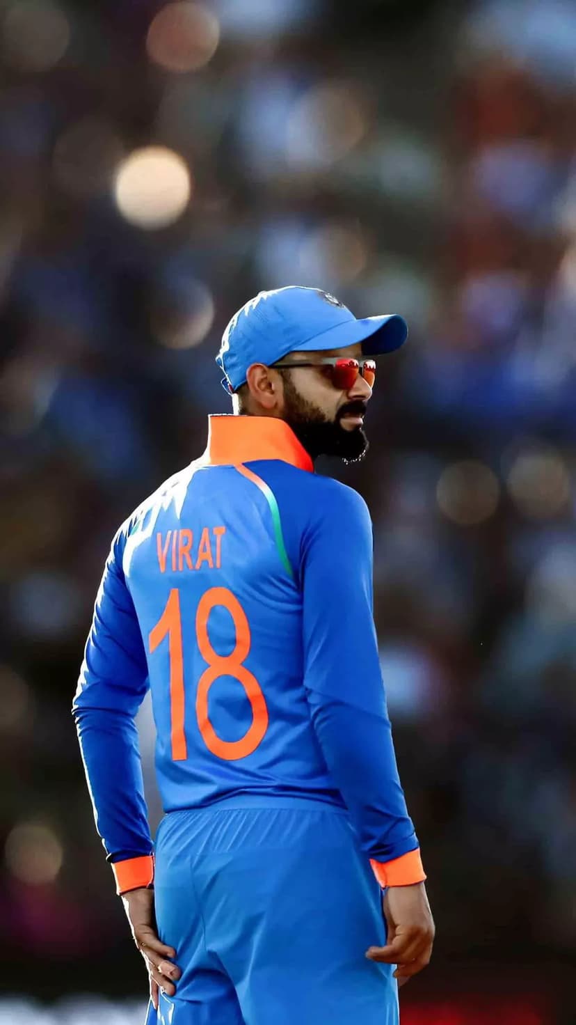 Virat Kohli in a blue India jersey with number 18, wearing sunglasses and a cap, standing in a blurred stadium.