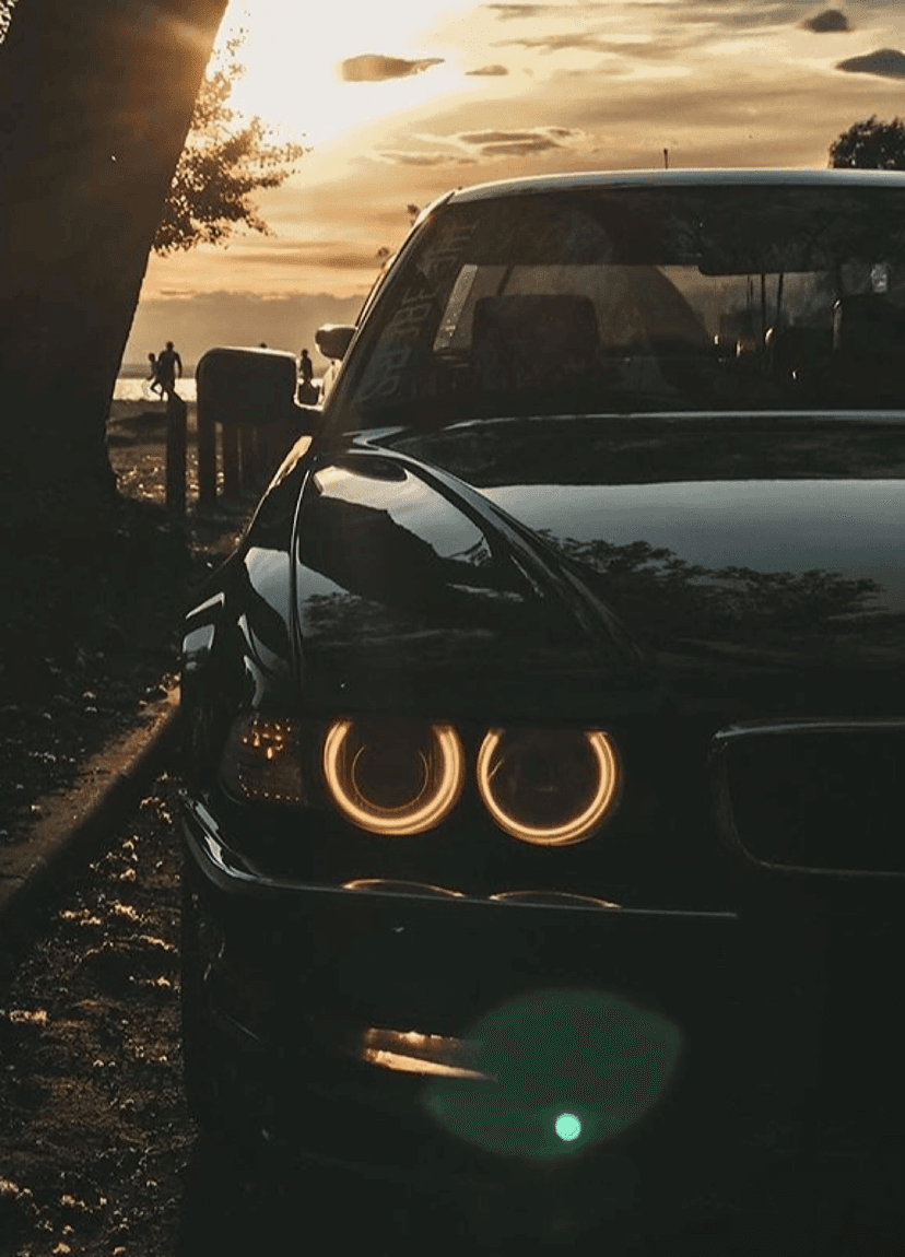 Illuminated BMW angel eye headlights reflect a golden sunset near a silhouetted tree and calm water at dusk.